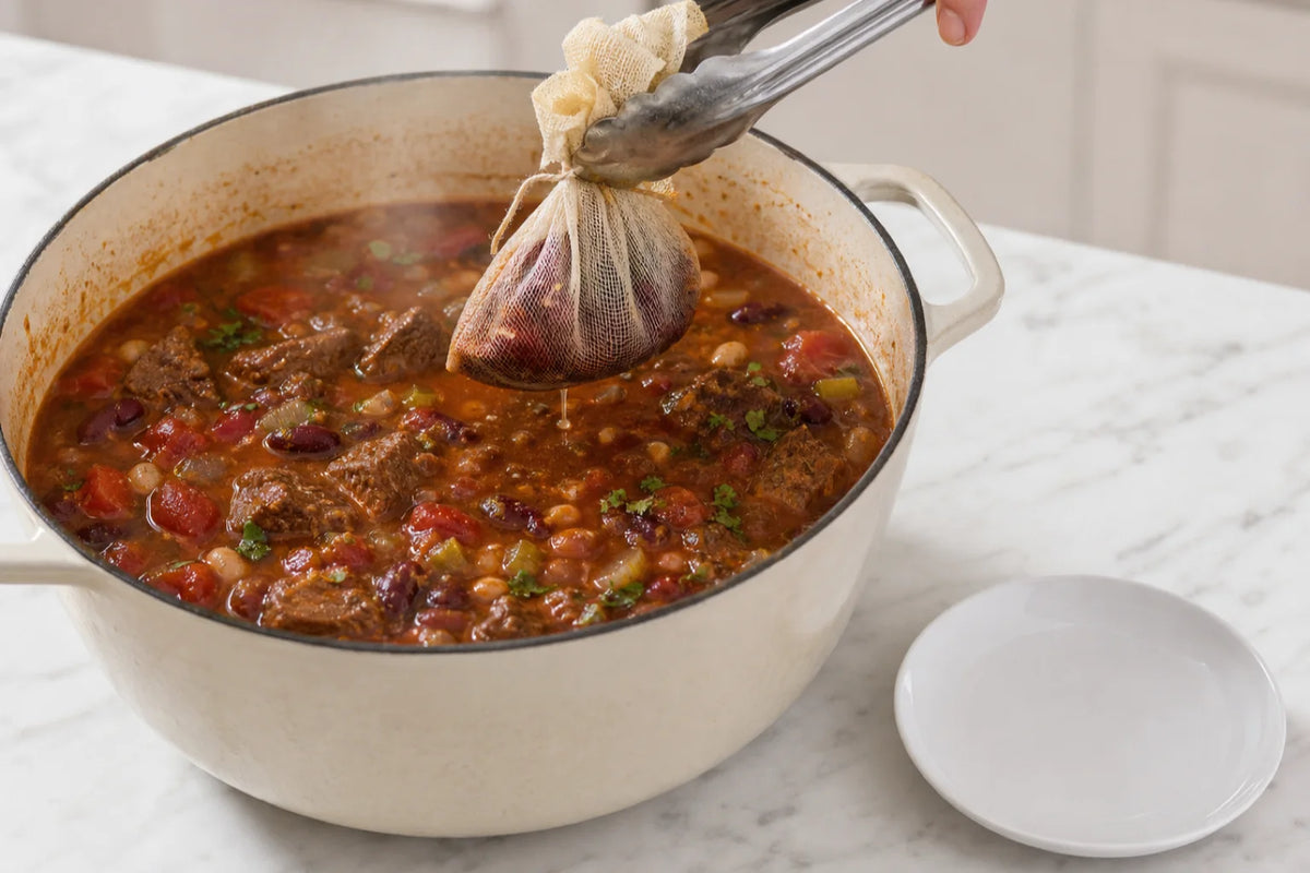 A cheesecloth pouch of dried chiles being lifted out of a pot of beef and bean chili with tongs, still dripping